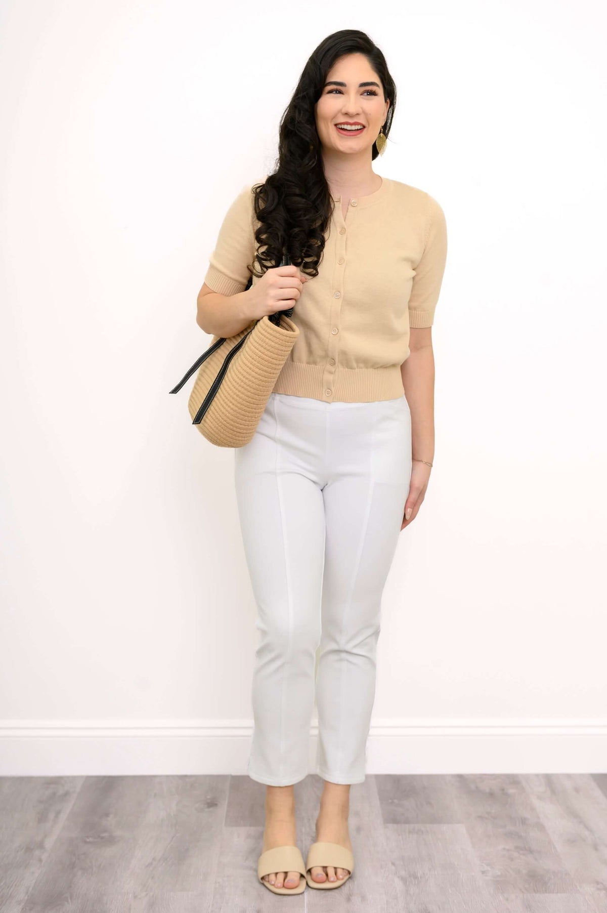 Woman in beige cardigan and white pants holding a tan bag, full-body front view