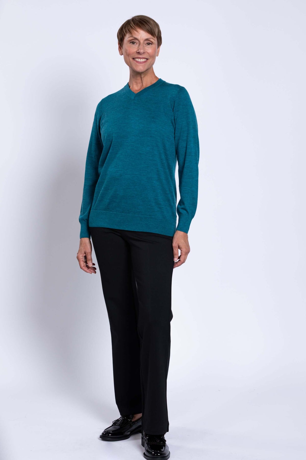 Full-body view of a woman in a teal V-neck sweater and black pants, standing against a white backdrop