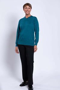 Full-body view of a woman in a teal V-neck sweater and black pants, standing against a white backdrop