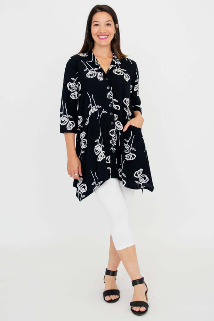 Full-body view of woman in black tunic with white floral print over white leggings, hand in pocket