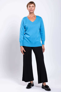 Full-length view of a woman in a blue V-neck sweater and black pants, facing forward