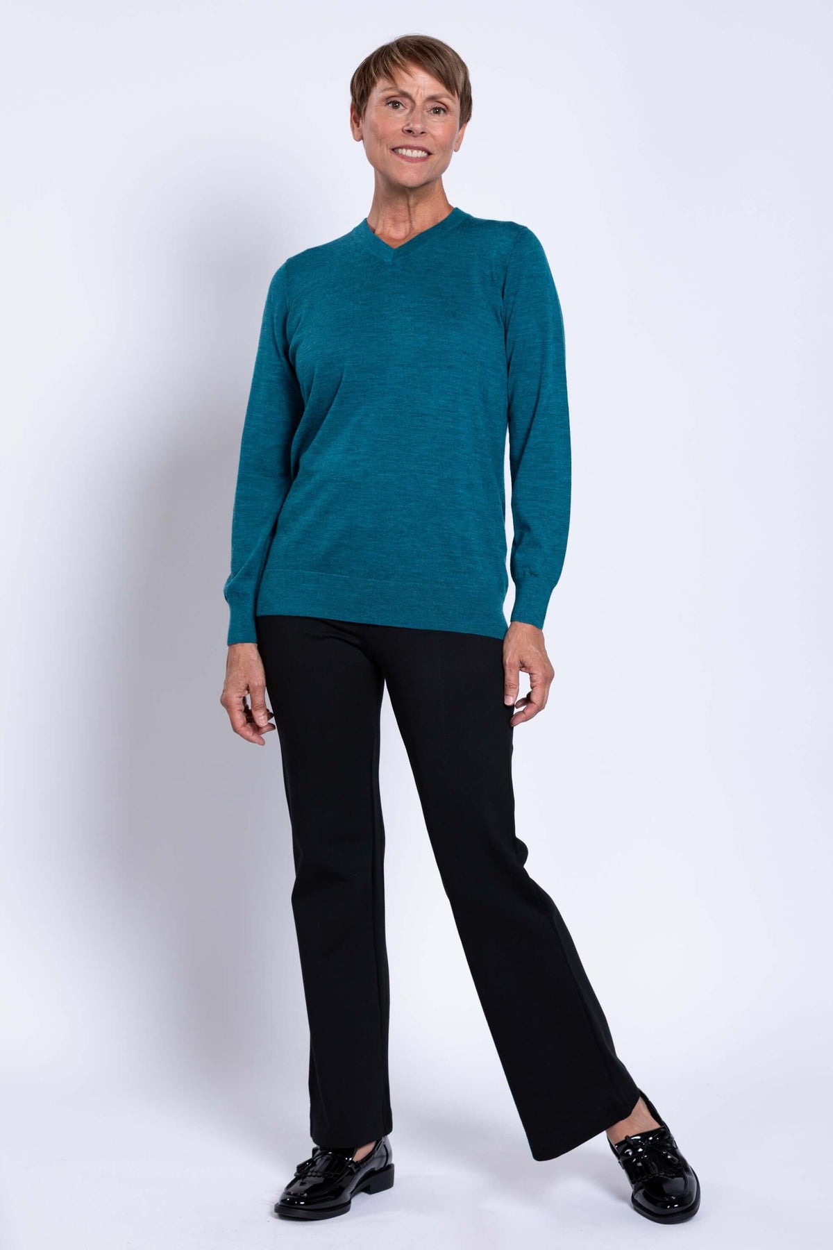 Full-length image of a woman in a teal V-neck sweater and black pants, smiling and standing naturally