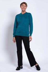 Full-length image of a woman in a teal V-neck sweater and black pants, smiling and standing naturally