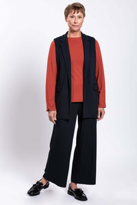 Model in black vest, matching pants, and rust long sleeve shirt, standing at an angle