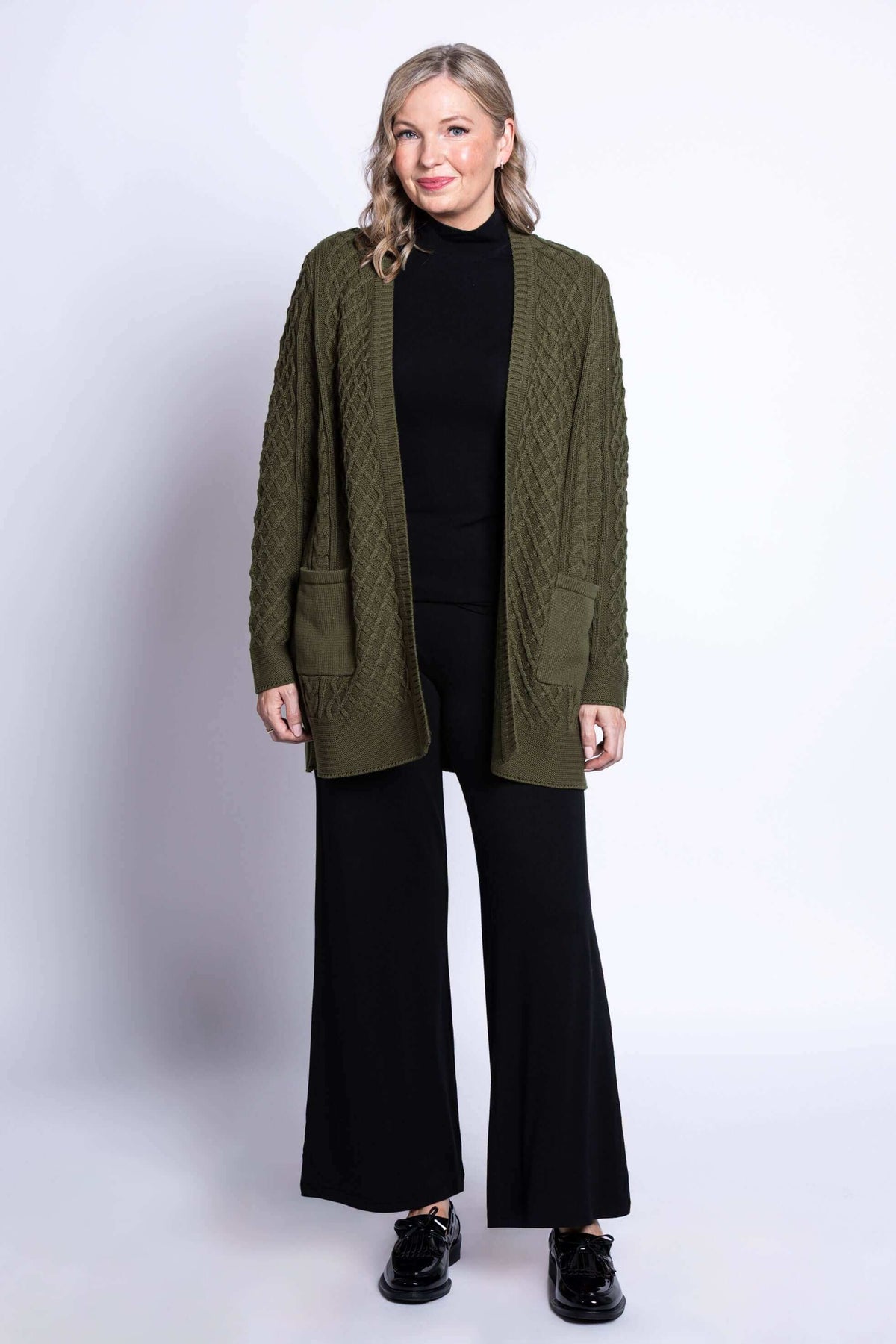 Full body view of a woman in an olive green cable knit cardigan and black pants