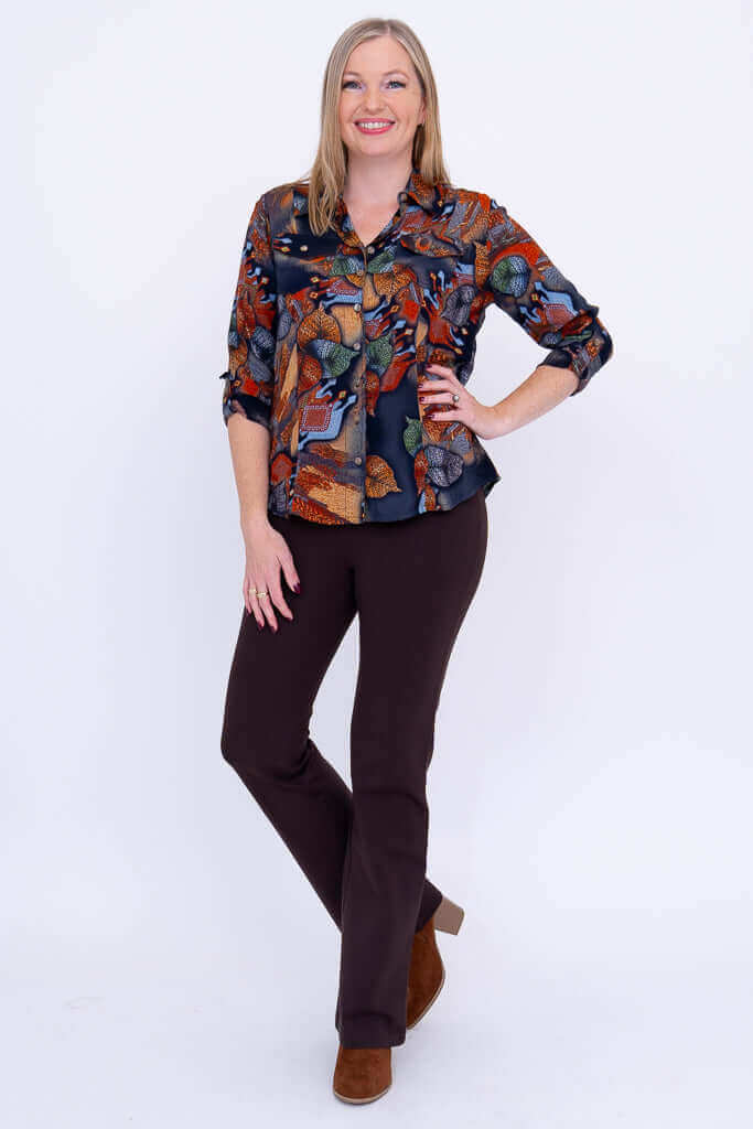 Full body shot of woman in botanical print blouse and brown pants standing against white background