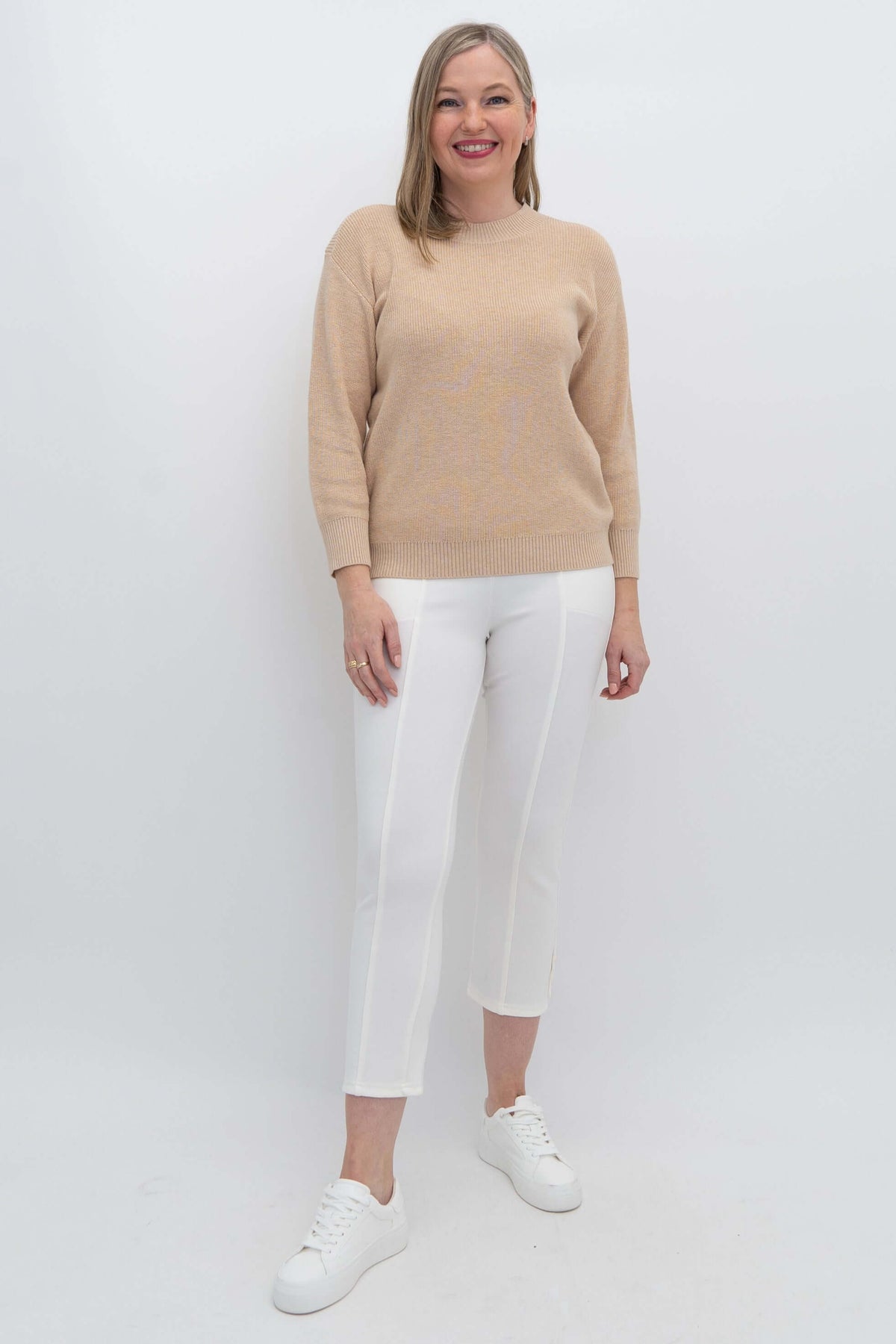 Full body view of woman in a beige crew neck sweater and white pants