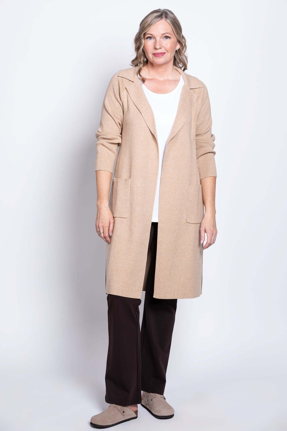 Full body view of a woman in a beige open front cotton jacket, white shirt, and brown pants