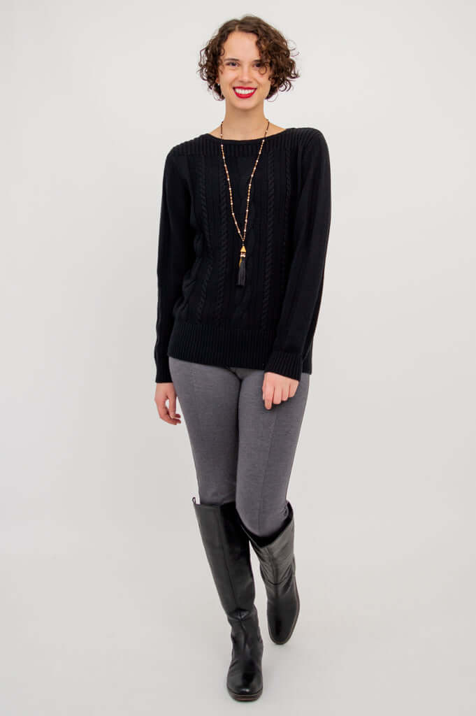 Full length view of a woman in a black cable knit sweater, grey pants, and black boots