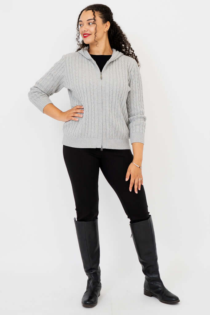 Full body view of woman in light grey cable knit hoodie sweater and black pants with boots