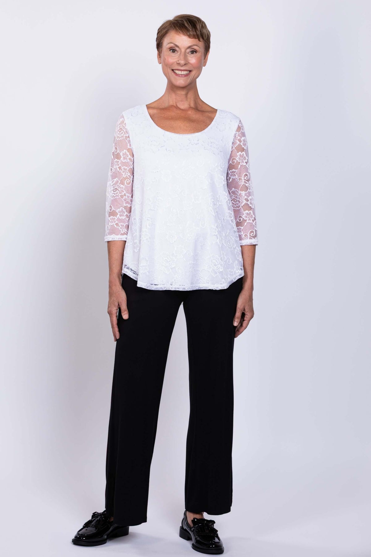 Full front view of a woman modeling a white lace overlay top with black pants and black shoes