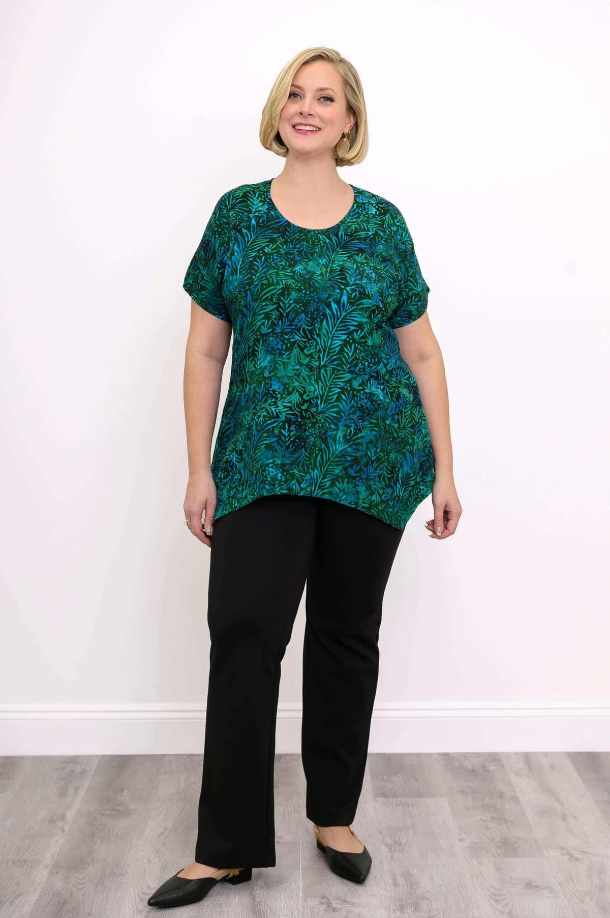 Full front view of woman standing in green and blue leafy print tunic top and black pants