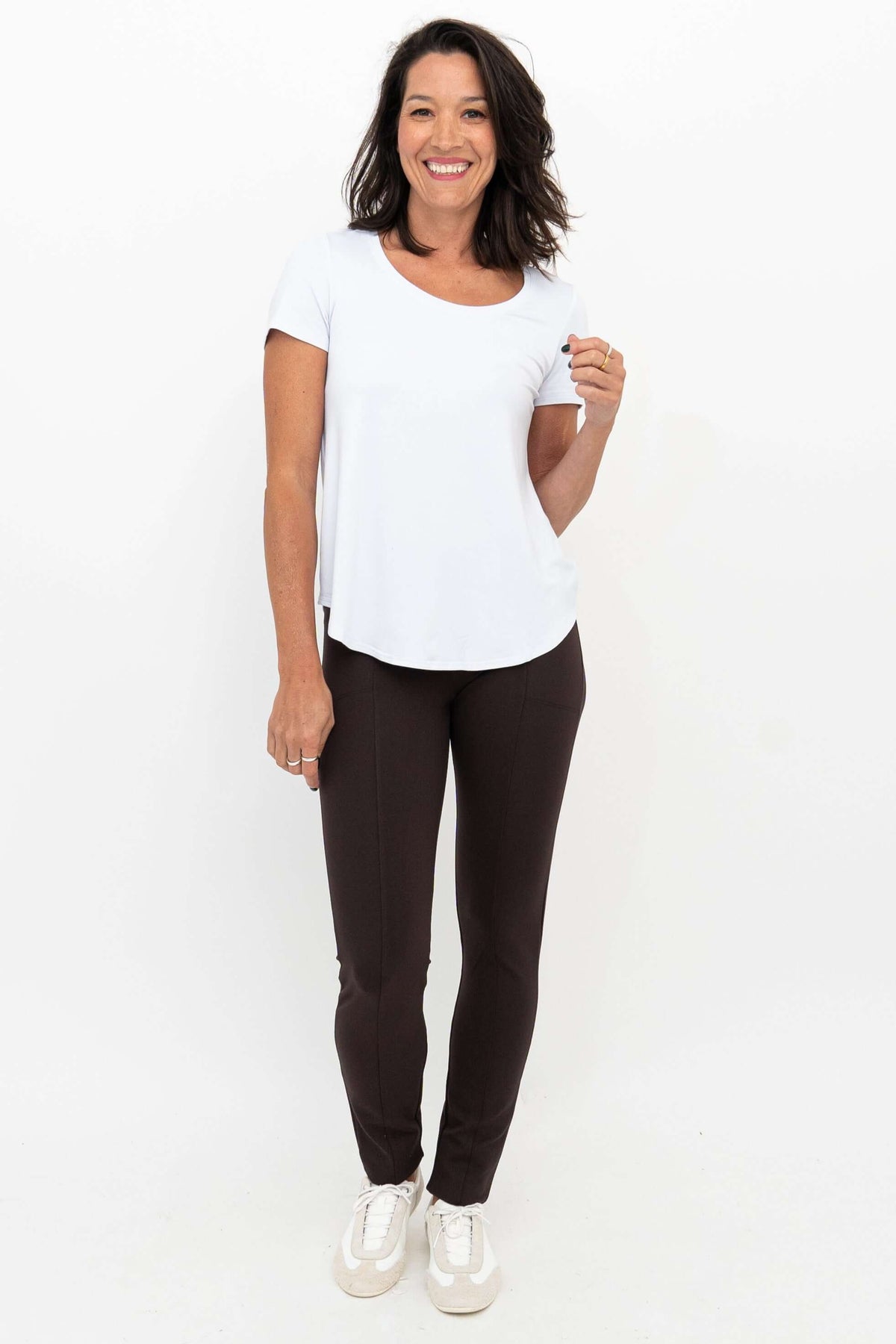 Woman smiling wearing Nadia slim-line brown pants and a white top, full front view