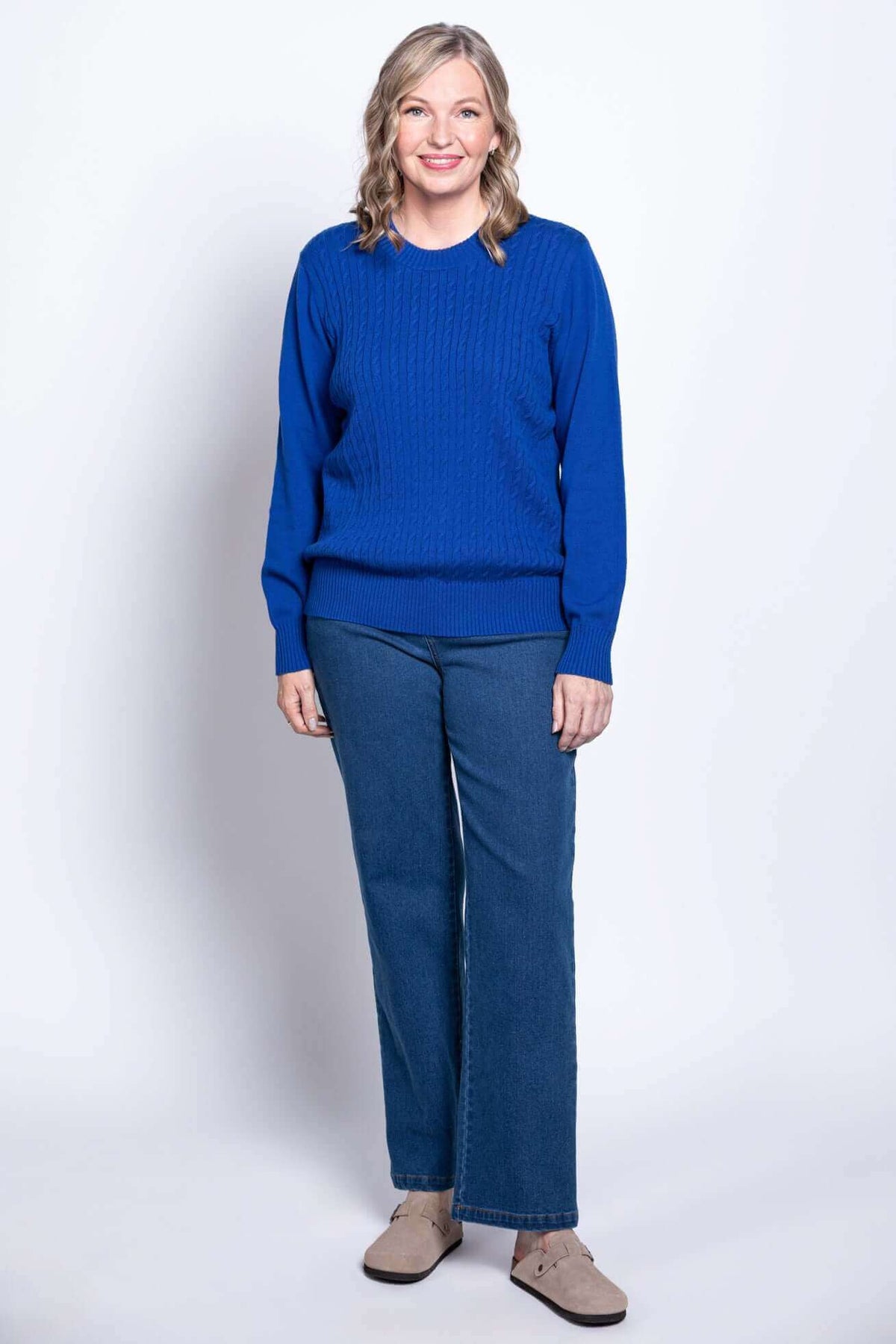 Full length view of a woman wearing a royal blue cable knit crewneck sweater with blue denim jeans.
