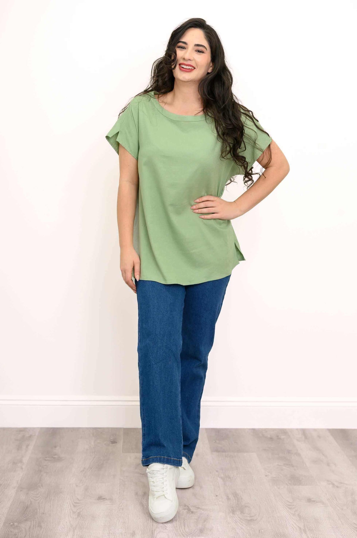 Full body view of a woman in a green linen tee paired with blue jeans and white sneakers