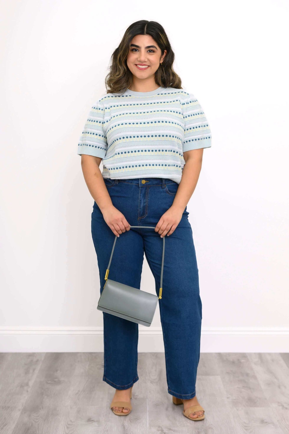 Woman in light blue sweater and blue jeans holding a gray purse, full length view