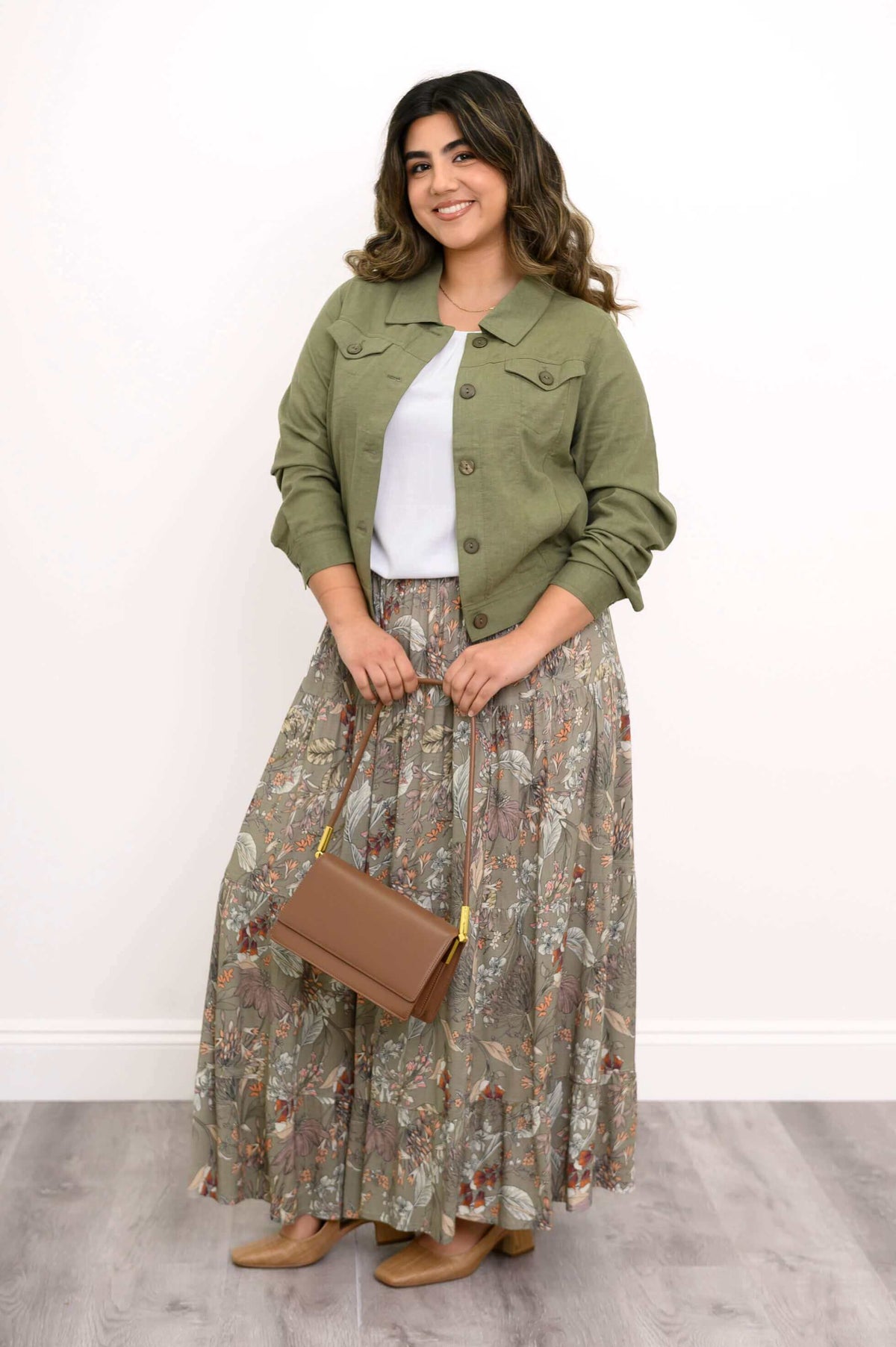 Woman posing in an olive green jacket, white blouse, floral skirt, and holding a brown handbag