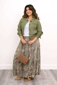 Woman posing in an olive green jacket, white blouse, floral skirt, and holding a brown handbag