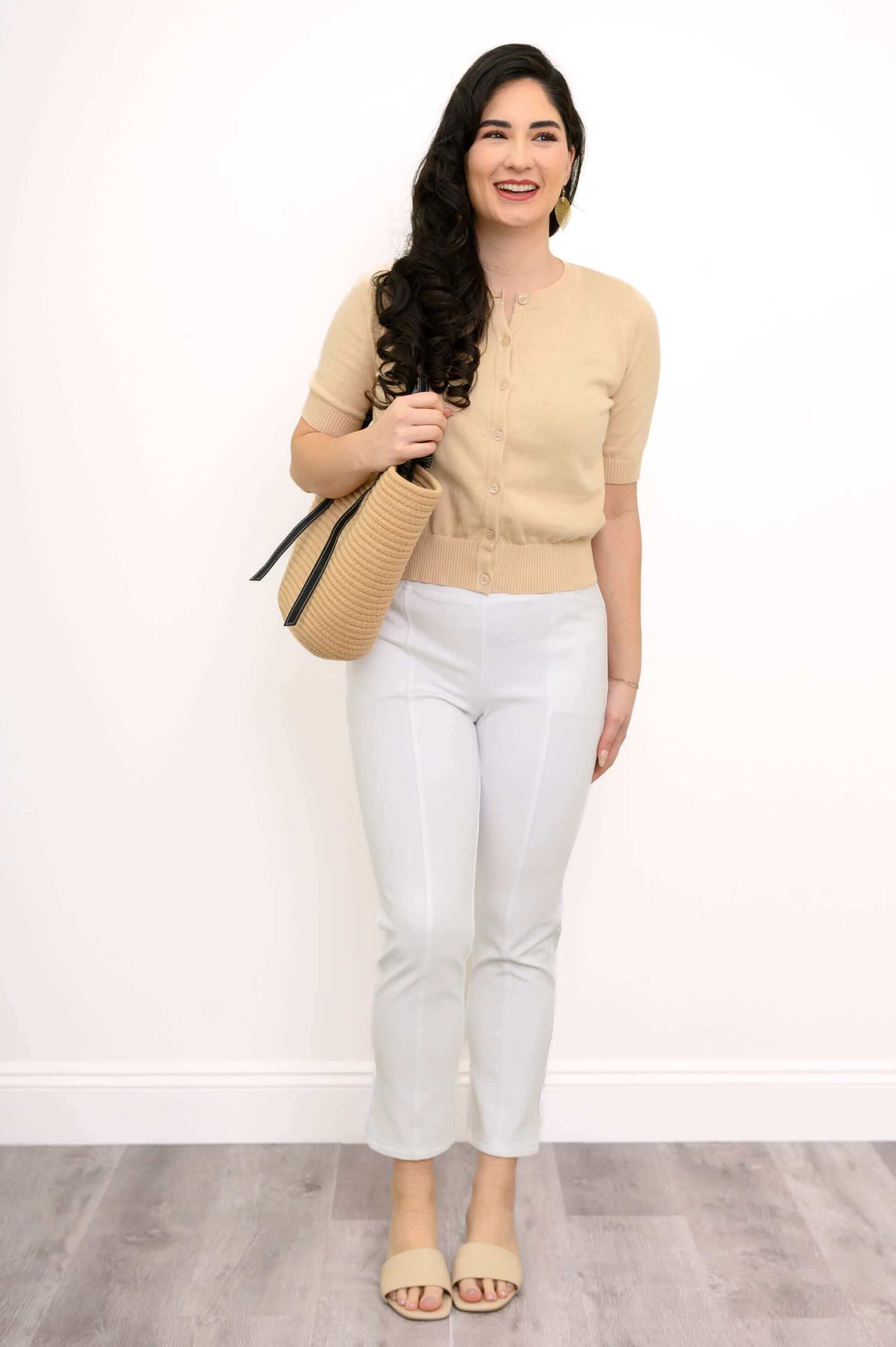 Woman in a beige cardigan, white pants, and beige sandals holding a tan bag