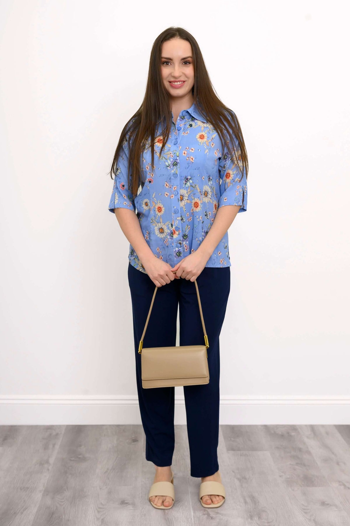 Full body view of a woman in a blue floral blouse with dark pants and beige accessories