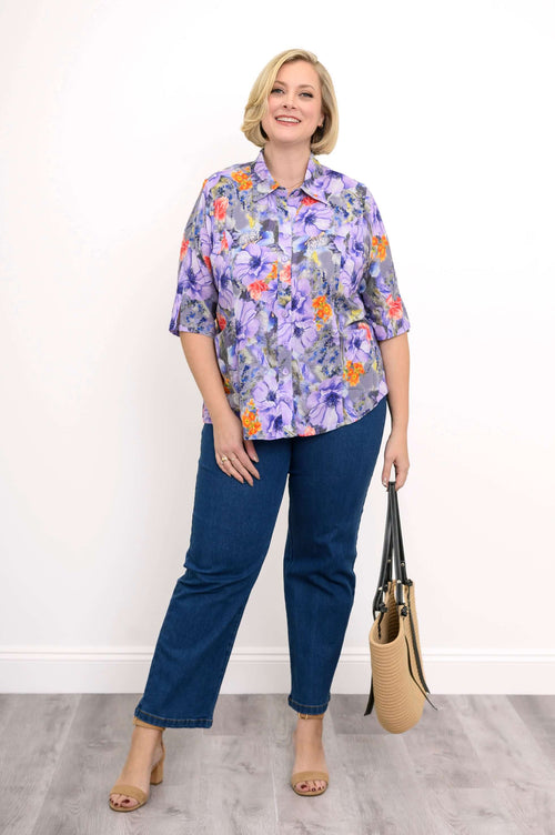 Full body view of a woman in a purple floral blouse, blue jeans, and tan heels holding a straw bag