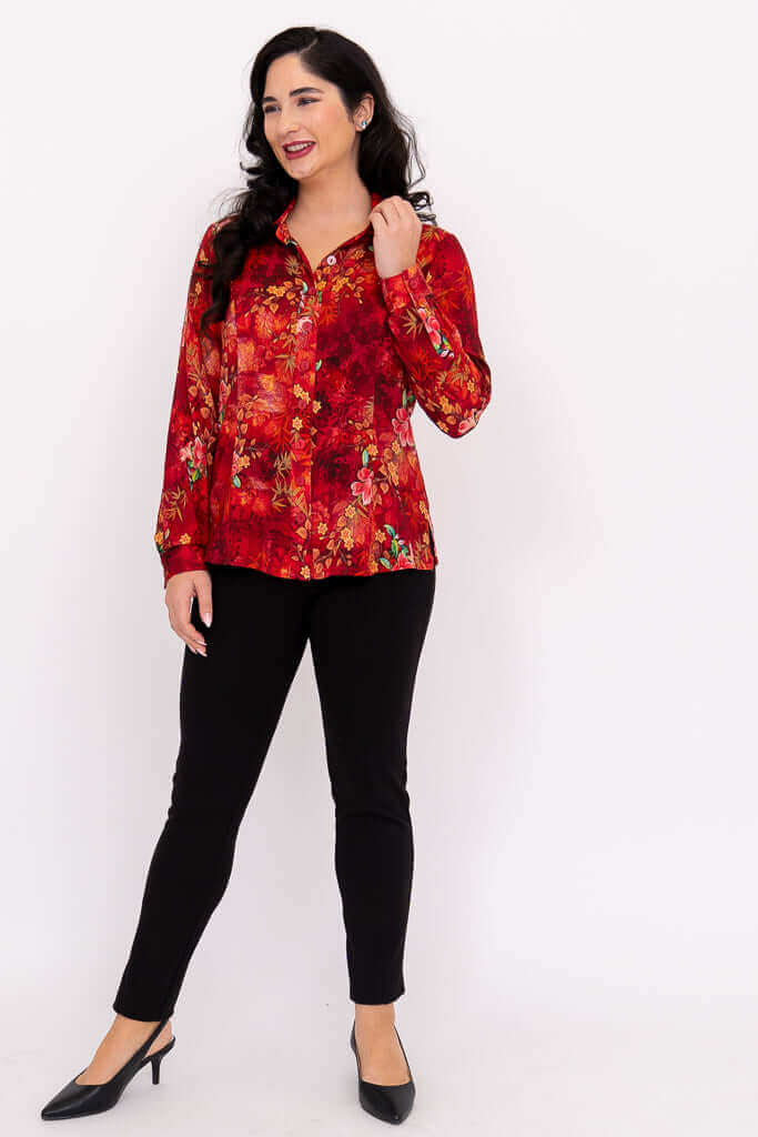 Full view of woman in red floral blouse and black pants, hand in hair, smiling