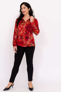 Full view of woman in red floral blouse and black pants, hand in hair, smiling