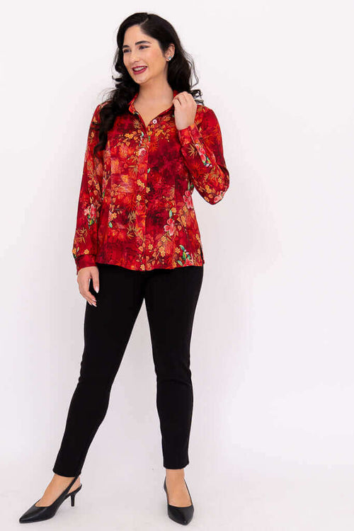 Full view of woman in red floral blouse and black pants, hand in hair, smiling