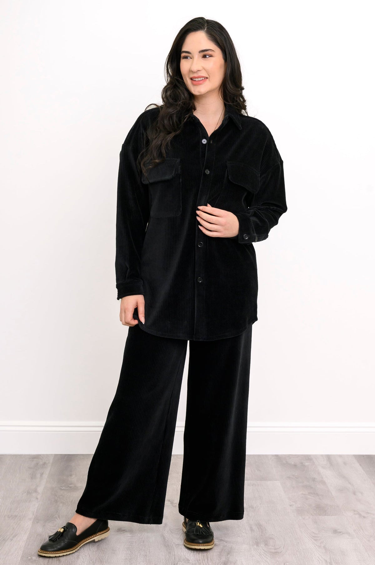A woman modeling a black velvet button-up shirt and matching wide-leg pants set, paired with black slip-on shoes, against a white wall and light gray wood floor.