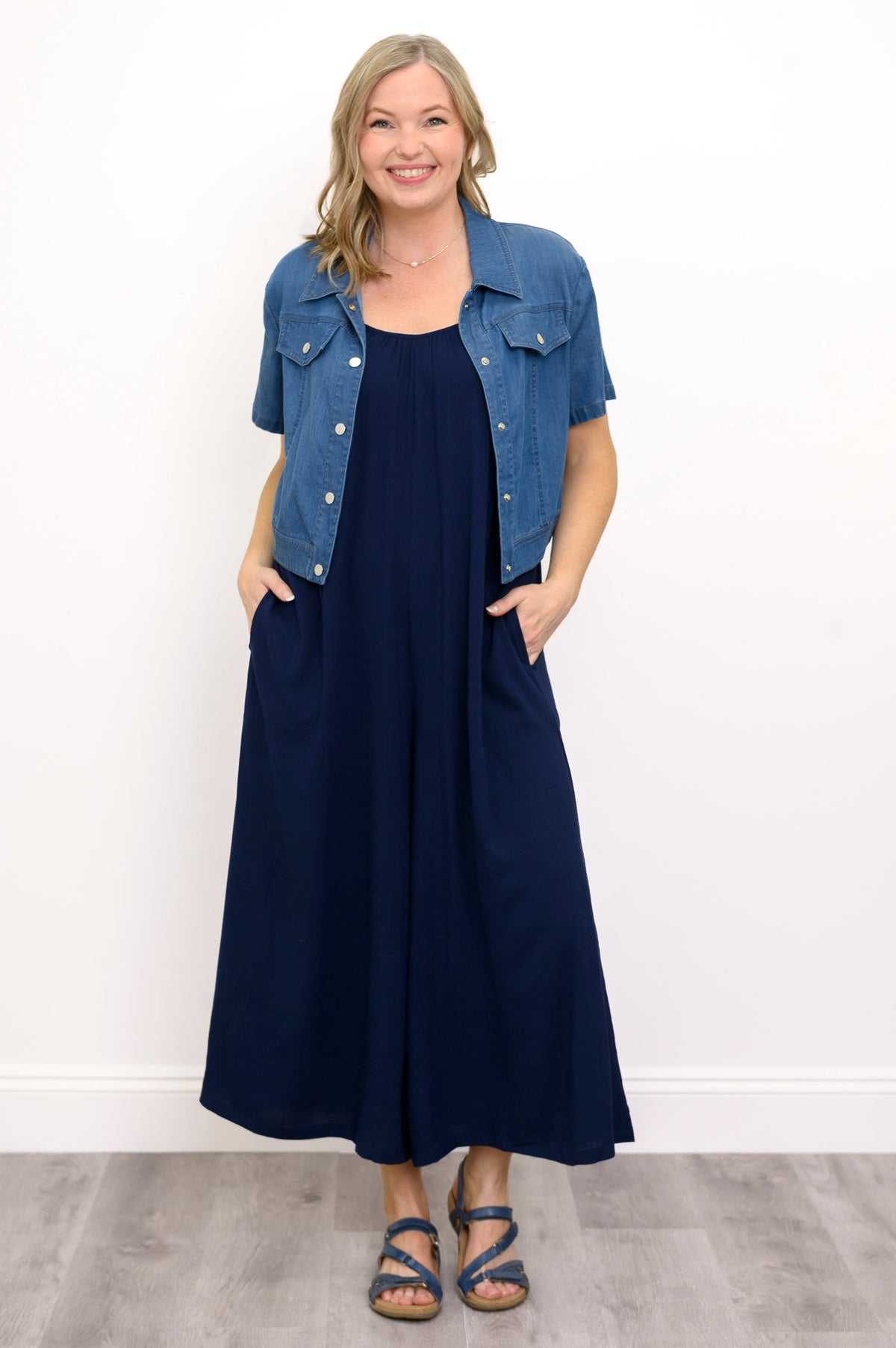 Front view, woman wearing blue jacket, navy blue jumpsuit, and sandals against a white background