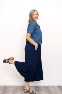 Side view, woman wearing blue jacket, navy blue jumpsuit, and sandals against a white background