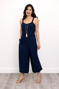 Woman wearing a navy blue jumpsuit, tassel necklace, and beige sandals against a white background, front view