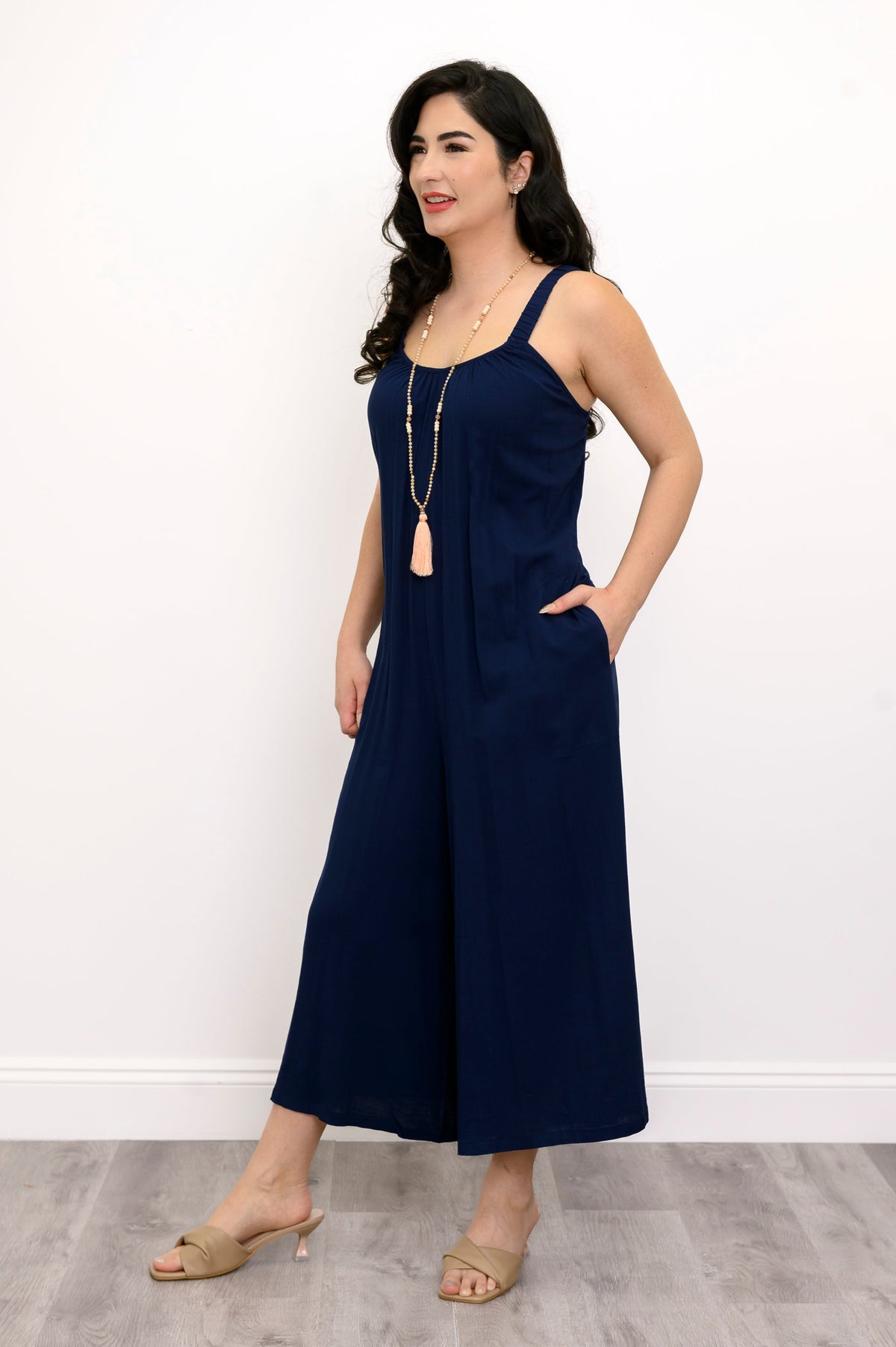 Woman wearing a navy blue jumpsuit, tassel necklace, and beige sandals against a white background, side view