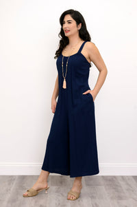 Woman wearing a navy blue jumpsuit, tassel necklace, and beige sandals against a white background, side view