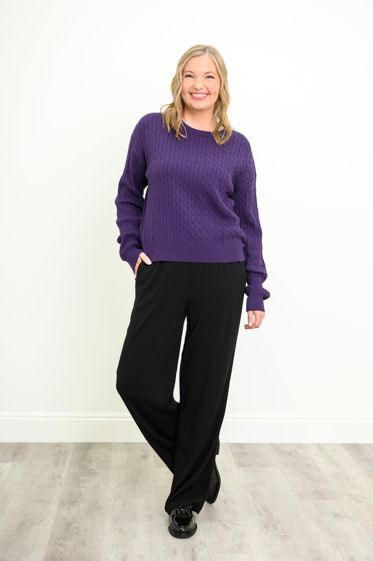 Smiling model in a cozy violet cable-knit pullover and black pants, posing naturally on a white background.