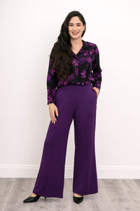 Full-length front view of a smiling woman wearing royale purple wide-leg pants paired with a patterned floral blouse.