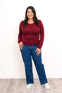 Deep maroon long-sleeve knit shirt featuring a flat finished hemline, styled simply with denim trousers, front view.