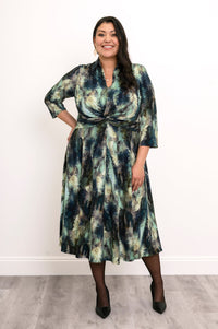 Plus-size model in a green forest print midi dress with a twisted waist, sheer black tights, and black heels.