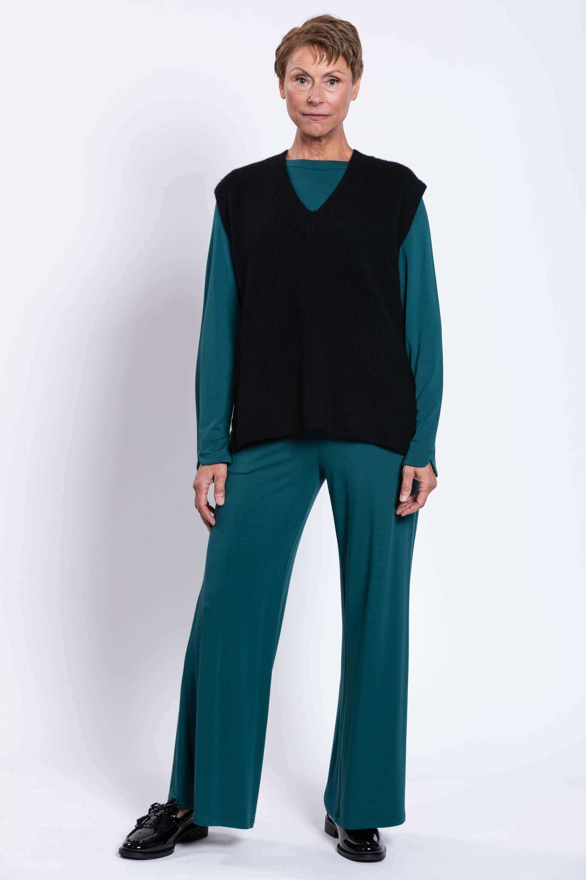 Model in black sweater vest with teal long sleeve shirt and flared trousers, front view