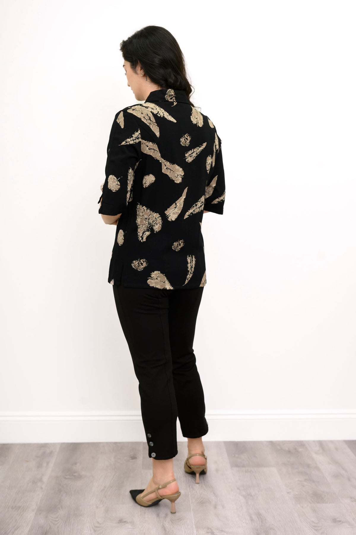 Woman wearing a black blouse with tan leaf pattern and black pants against a white background, black view