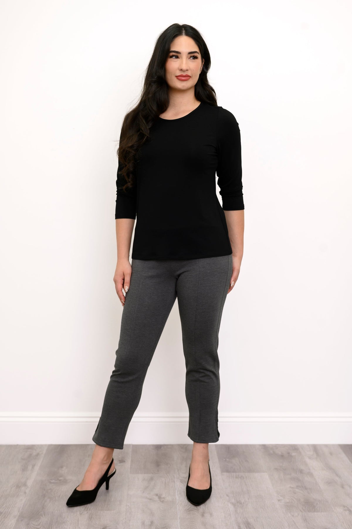 Full body front view of woman wearing black long sleeve shirt, grey pants, and black heels