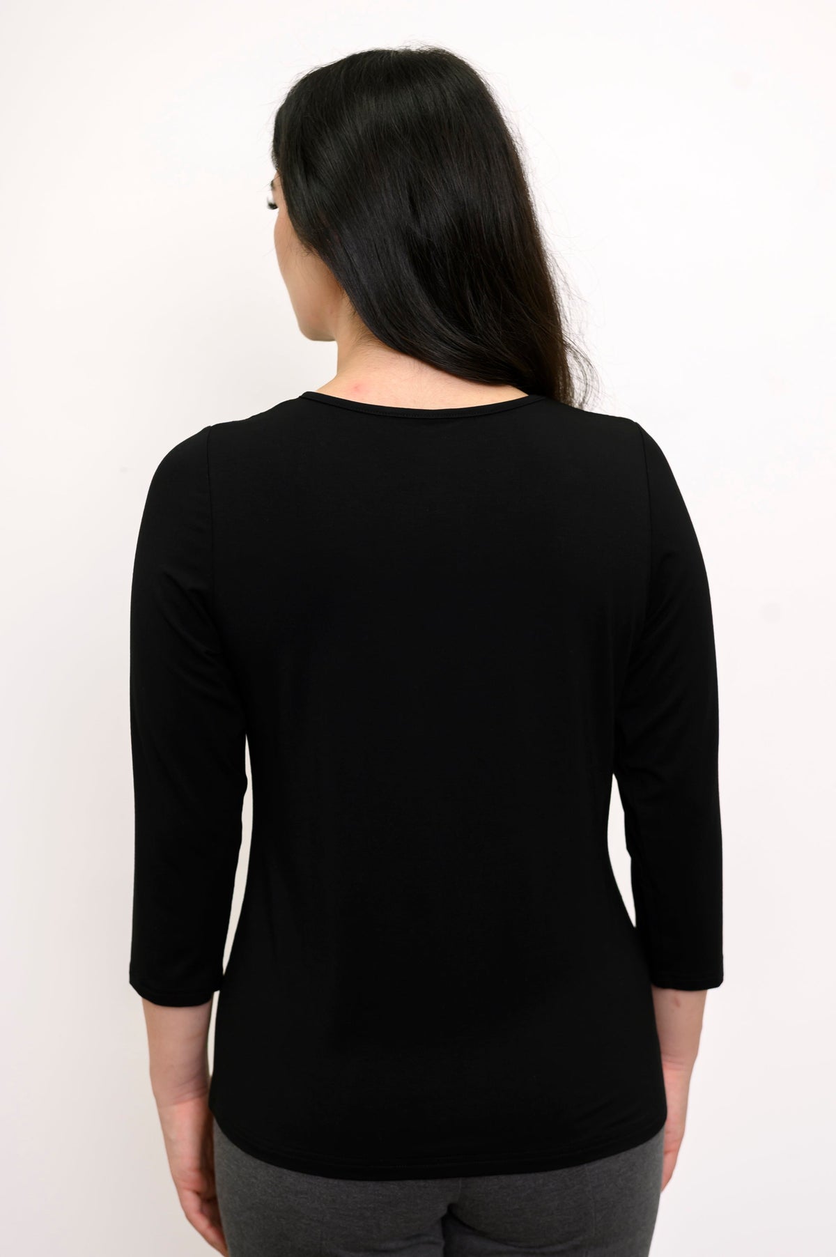 Back view of woman wearing black 3/4 sleeve top with slim fit grey pants against a white background