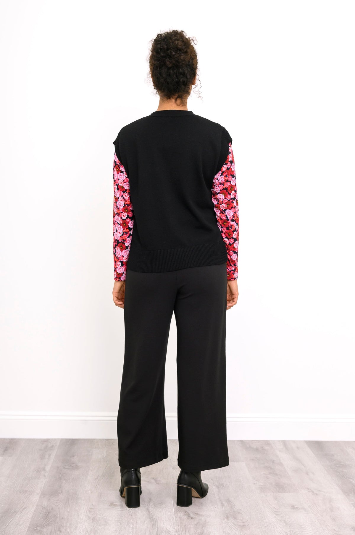 Back view of a women's V-neck black knit vest, styled with a pink patterned shirt and black trousers.