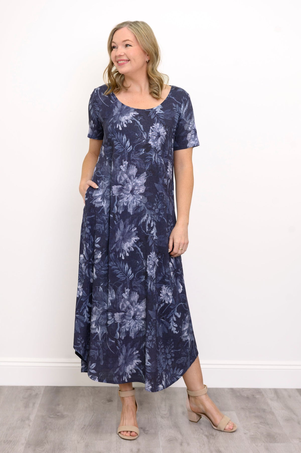 Front view of woman wearing long navy blue floral dress with pockets and sandals on a white background