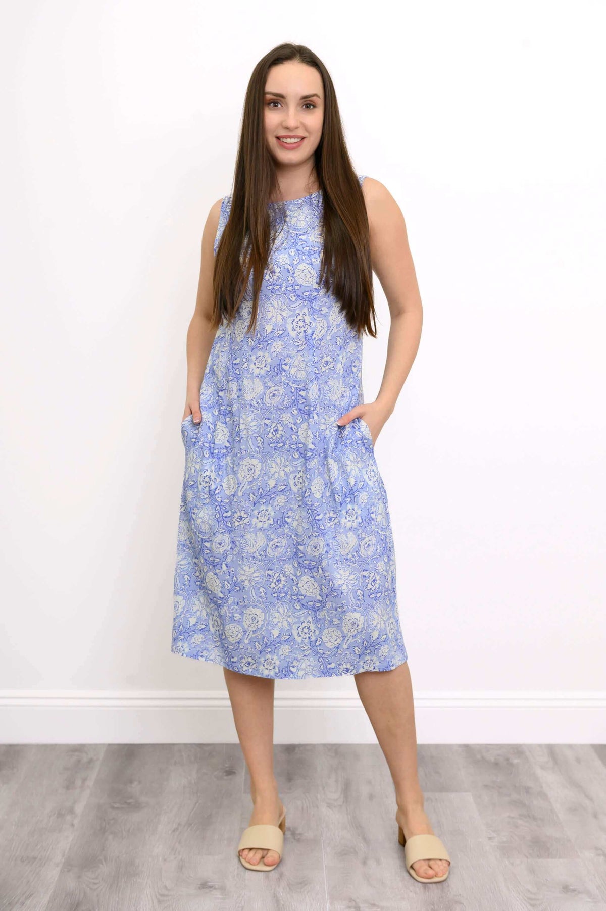Full body image of model wearing long sleeveless blue floral dress with her hands in her pockets