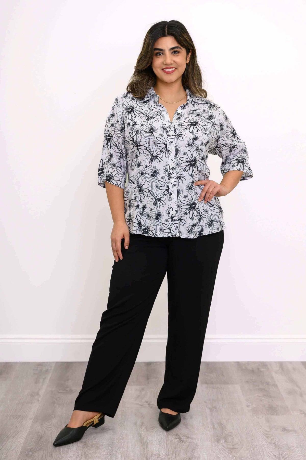 Model standing, hand on hip, in a black and white floral blouse and black pants