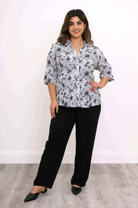 Model standing, hand on hip, in a black and white floral blouse and black pants