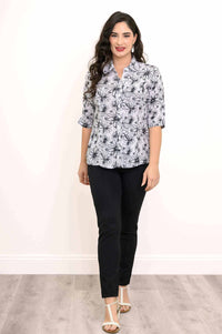 Model walking forward in a floral bamboo linen blouse with black pants and white sandals