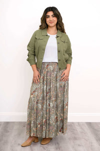 Woman in olive green jacket and floral skirt standing with hands at sides