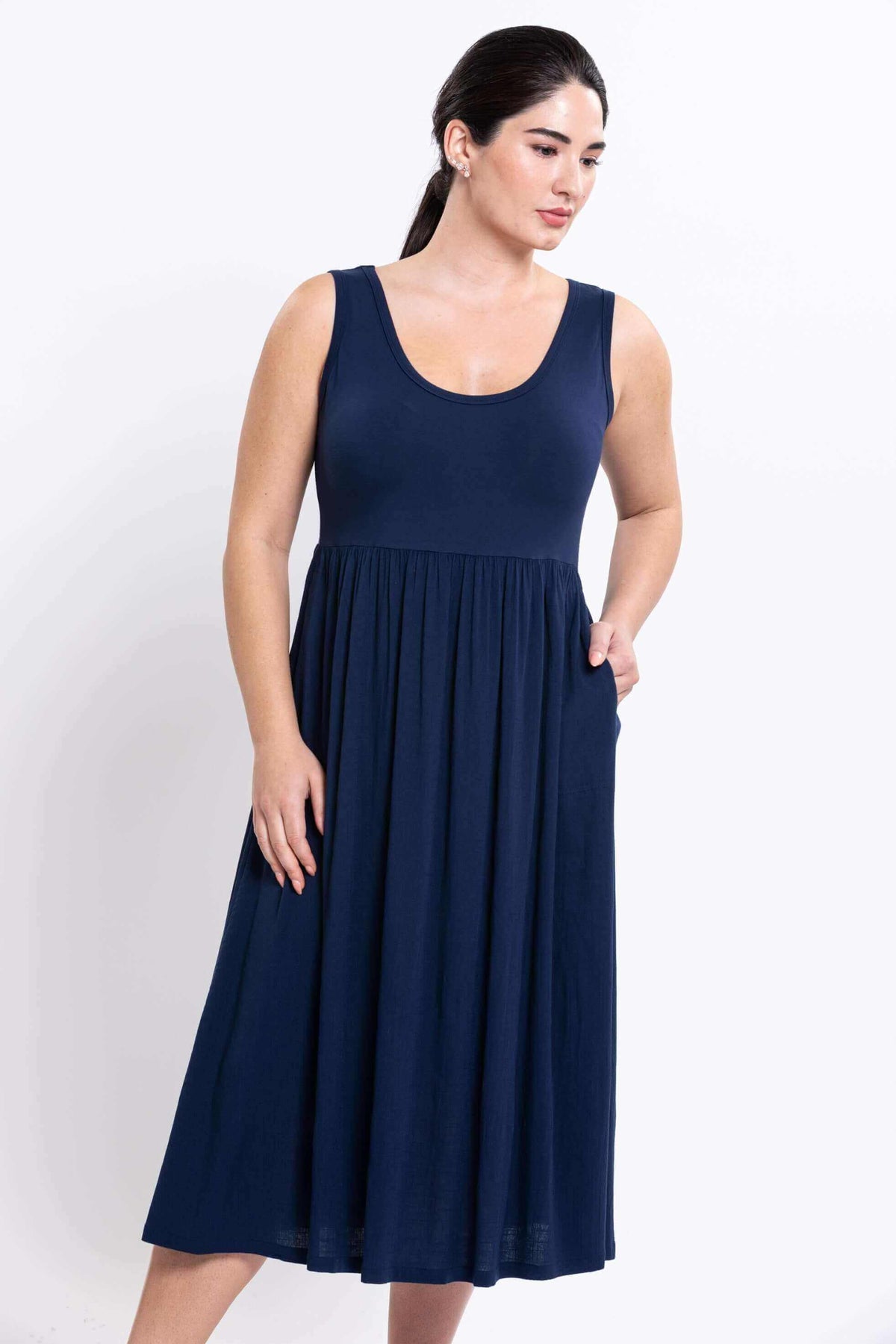 Woman in navy empire waist midi dress, left hand in pocket, looking down, front view